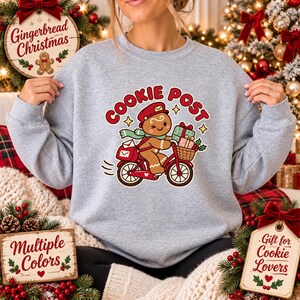 May include: A light gray crewneck sweatshirt featuring a graphic of a gingerbread person wearing a red mail carrier hat and a green scarf, riding a red bicycle. The text above reads COOKIE POST in red letters, with small stars and gift boxes.
