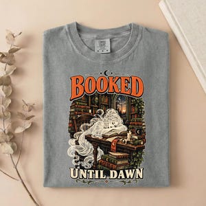 May include: A grey t-shirt featuring a graphic print of a person with long flowing hair resting their head on an open book at a desk. The text reads BOOKED at the top and UNTIL DAWN at the bottom, surrounded by stacks of books and a window.