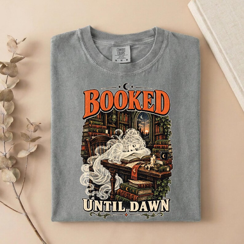 May include: A grey t-shirt featuring a graphic print of a person with long flowing hair resting their head on an open book at a desk. The text reads BOOKED at the top and UNTIL DAWN at the bottom, surrounded by stacks of books and a window.