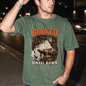 May include: A sage green short-sleeve t-shirt featuring a graphic print of a ghost reading a book at a desk in a library. The text BOOKED is printed in orange above the illustration, and the text UNTIL DAWN is printed in white below the illustration.