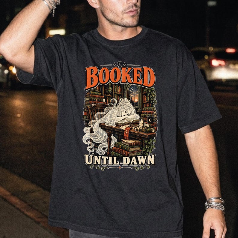May include: A black short-sleeve t-shirt featuring a graphic print of a ghost sleeping at a desk in a library. The text "BOOKED" appears in orange above the illustration, and "UNTIL DAWN" is printed in white below the image of books and candles.