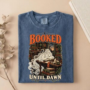 May include: A blue t-shirt featuring a graphic print of a ghost reading a book at a desk surrounded by bookshelves. The text reads BOOKED at the top and UNTIL DAWN at the bottom. The design includes stacks of books, a candle, and a crescent moon.