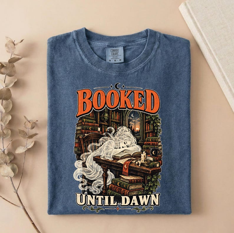 May include: A blue t-shirt featuring a graphic print of a ghost reading a book at a desk surrounded by bookshelves. The text reads BOOKED at the top and UNTIL DAWN at the bottom. The design includes stacks of books, a candle, and a crescent moon.