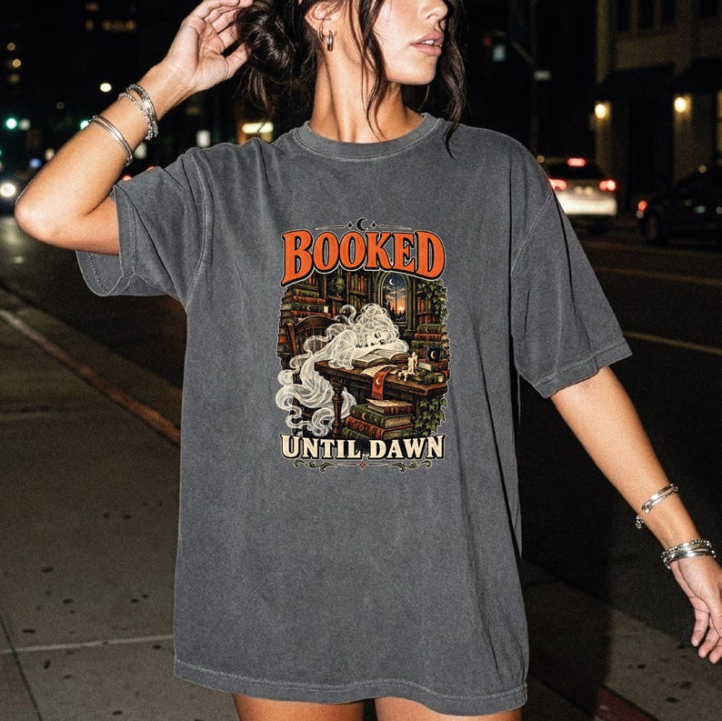 May include: A charcoal gray short-sleeve t-shirt featuring a graphic print of a person reading at a desk surrounded by bookshelves. The text BOOKED is printed in orange above the illustration, and UNTIL DAWN is printed in white below the image.