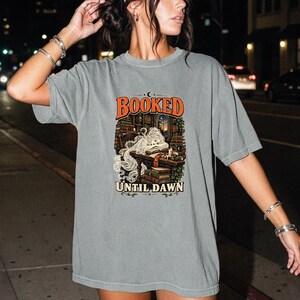 May include: A grey short-sleeve t-shirt featuring a graphic print of a person sleeping at a desk in a library with stacks of books. The text "BOOKED" appears in orange above the illustration, and "UNTIL DAWN" is printed in white at the bottom.