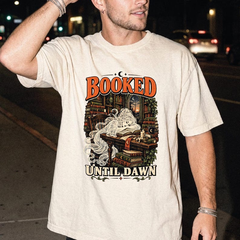 May include: A beige short-sleeve t-shirt featuring a graphic print of a person sleeping at a desk in a library. The text "BOOKED" appears in orange above the illustration, and "UNTIL DAWN" is printed in black below the scene with books and candles.