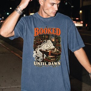 May include: A blue short-sleeve t-shirt featuring a graphic print of a ghost reading at a desk in a library. The text "BOOKED" appears in orange above the illustration, and the text "UNTIL DAWN" appears in cream below the illustration.
