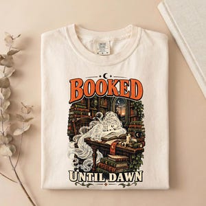 May include: A cream-colored t-shirt featuring a graphic print with the text BOOKED UNTIL DAWN. The illustration depicts a translucent figure resting at a wooden desk in a library filled with books, a candle, and a window showing a sunset.