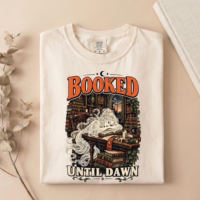 May include: A cream-colored t-shirt featuring a graphic print with the text BOOKED UNTIL DAWN. The illustration depicts a translucent figure resting at a wooden desk in a library filled with books, a candle, and a window showing a sunset.