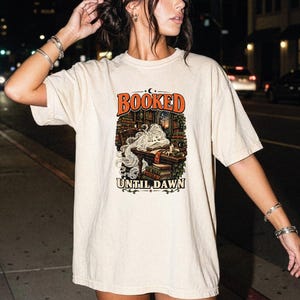 May include: A beige short-sleeve t-shirt featuring a graphic print of a person sleeping at a desk in a library surrounded by books. The text "BOOKED" appears in orange above the illustration, and "UNTIL DAWN" is printed in black below the image.