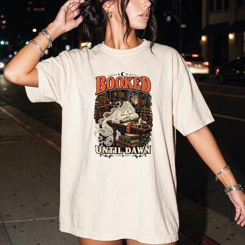 May include: A beige short-sleeve t-shirt featuring a graphic print of a person sleeping at a desk in a library surrounded by books. The text "BOOKED" appears in orange above the illustration, and "UNTIL DAWN" is printed in black below the image.