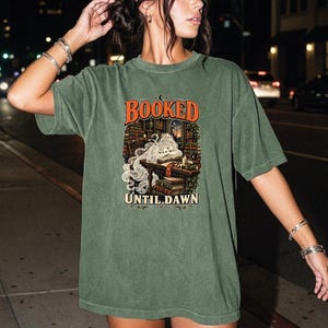 May include: An olive green short-sleeve t-shirt featuring a graphic print of a person sleeping at a desk surrounded by stacks of books. The text "BOOKED" appears in orange above the illustration, and "UNTIL DAWN" is printed in white below it.