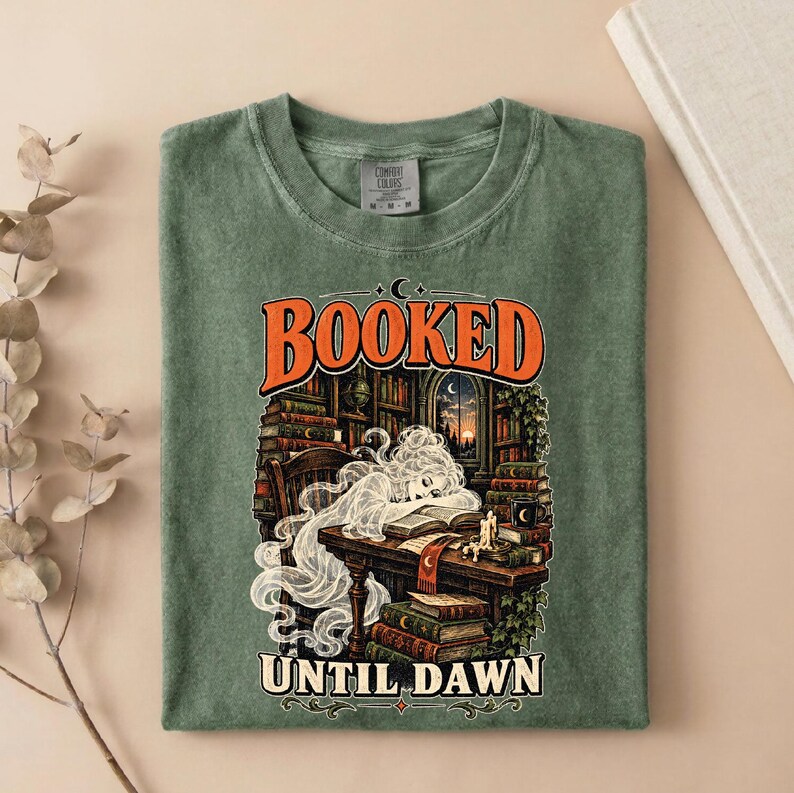 May include: A sage green t-shirt featuring a graphic print of a ghost reading a book at a desk in a library. The text "BOOKED" appears in orange above the illustration, and "UNTIL DAWN" is printed in white below. The shirt has a crew neckline.
