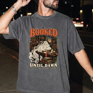 May include: A charcoal gray short-sleeve t-shirt featuring a graphic print of a ghost reading at a desk in a library. The text "BOOKED" appears in orange above the illustration, and "UNTIL DAWN" is printed in cream below the image of books.