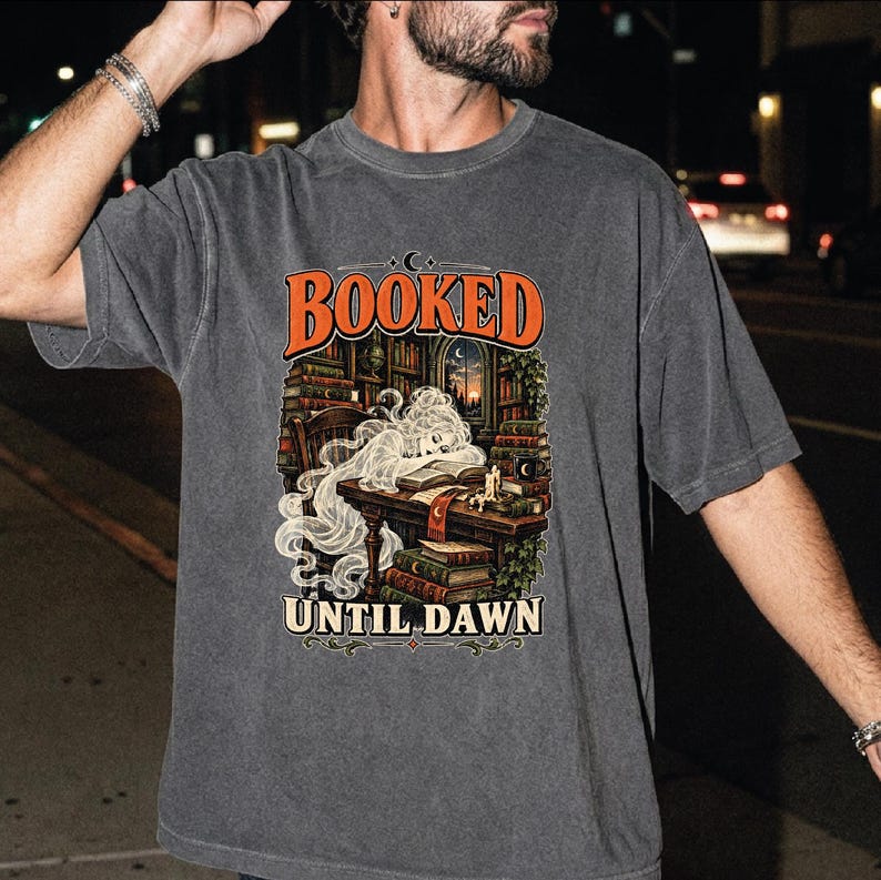 May include: A charcoal gray short-sleeve t-shirt featuring a graphic print of a ghost reading at a desk in a library. The text "BOOKED" appears in orange above the illustration, and "UNTIL DAWN" is printed in cream below the image of books.