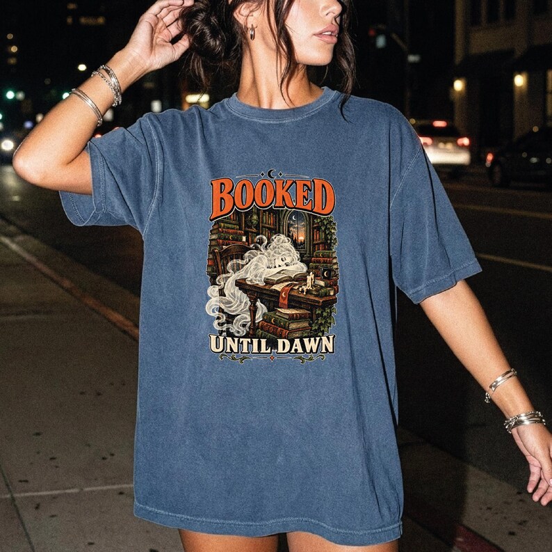 May include: A blue short-sleeve t-shirt featuring a graphic print of a person sleeping at a desk in a library with stacks of books and a candle. The text "BOOKED" appears in orange above the illustration and "UNTIL DAWN" in white below it.