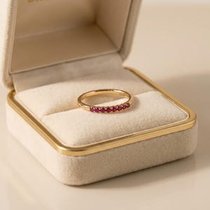 May include: A gold band ring with a row of small, vibrant red gemstones. The ring is displayed inside a cream-colored jewelry box with a gold trim, set against a soft, neutral background. The ring is a delicate piece of jewelry.
