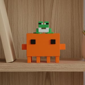 Peut inclure: Une figurine de créature orange au style pixel-art, composée de blocs, avec quatre pattes, deux bras latéraux et deux yeux carrés noirs. Une petite figurine de grenouille pixelisée verte et blanche est posée sur la tête de la créature orange.