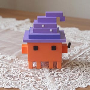 May include: A quirky, orange, pixelated figure wearing a purple witch's hat and black square eyes. The figure has small, blocky legs and arms. The object is on a white lace tablecloth.