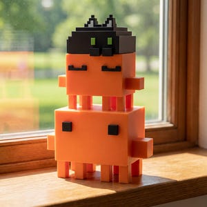 May include: A bright orange, pixelated cat ornament with a black head and square eyes. The cat is constructed from stacked blocks and is positioned on a wooden windowsill. The background features a blurred green garden.