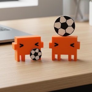 May include: Two orange block-shaped figures with simple black facial features. One figure has a small soccer ball attached to its side, while the other figure has a larger soccer ball balanced on its top. Both figures have a smooth, matte finish.