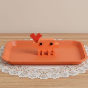 Puede incluir: Una bandeja rectangular de color naranja con una figura de criatura naranja pixelada que tiene dos peque&ntilde;os ojos cuadrados negros y una forma de coraz&oacute;n rojo pixelado unida a la cabeza. La bandeja tiene esquinas redondeadas y un acabado liso.