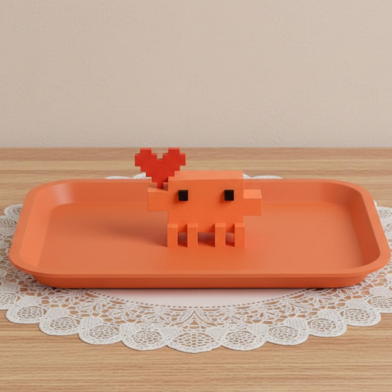 Puede incluir: Una bandeja rectangular de color naranja con una figura de criatura naranja pixelada que tiene dos peque&ntilde;os ojos cuadrados negros y una forma de coraz&oacute;n rojo pixelado unida a la cabeza. La bandeja tiene esquinas redondeadas y un acabado liso.