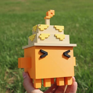 Puede incluir: Figura naranja y amarilla impresa en 3D con un diseño en bloques. La figura tiene un cuerpo cuadrado, ojos negros en forma de V y una estructura escalonada en la parte superior, que se asemeja a un pastel. Fondo verde.