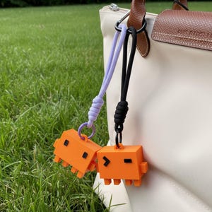 May include: Two orange pixelated creature keychains with black accents and attached to a purple and black lanyard. The keychains are hanging from a white purse with a brown leather handle, set against a backdrop of green grass.