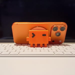 以下が含まれることがあります： オレンジ色のスマートフォンと、お揃いのオレンジ色の携帯電話ケース、小さなオレンジ色のロボット型携帯電話スタンド。 電話とスタンドは白いキーボードの上に置かれています。