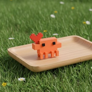 Puede incluir: Una peque&ntilde;a figura de cangrejo naranja pixelado con una pinza roja en forma de coraz&oacute;n. El cangrejo tiene un dise&ntilde;o geom&eacute;trico de bloques con dos peque&ntilde;os ojos cuadrados negros y varias patas, sobre una bandeja rectangular de madera clara.