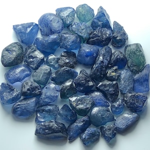 Puede incluir: Una colección de piedras preciosas de zafiro en bruto y sin pulir en varios tonos de azul profundo, azul marino y azul claro. Estas piedras naturales presentan formas irregulares y dentadas con superficies de translúcidas a opacas y una textura cristalina.