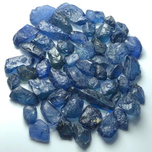 Puede incluir: Una colección de piedras preciosas de zafiro azul en bruto y sin pulir de diversas formas y tamaños. Las piedras presentan una gama de tonos desde el azul profundo hasta el azul claro con texturas translúcidas y opacas, facetas naturales y bordes irregulares.