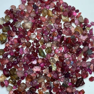 Puede incluir: Una colección de piedras preciosas en bruto sueltas en varios tonos de rosa, rojo, morado y ámbar. Estas pequeñas piedras de formas irregulares presentan texturas facetadas y rugosas, mostrando una mezcla de acabados translúcidos y opacos.