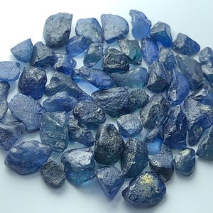 Könnte beinhalten: Eine Ansammlung von rohen, ungeschliffenen blauen Saphiredelsteinen in verschiedenen Formen und Größen. Die Steine zeigen eine Reihe von tiefblauen bis hellblauen Farbtönen, wobei einige Stücke durchscheinend und andere undurchsichtig sind.