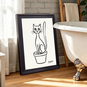 May include: A framed black and white line drawing print featuring a cat sitting inside a flower pot. The cat has large eyes, pointed ears, and a long upright tail. The word Picasso is written in black cursive script in the bottom right corner.