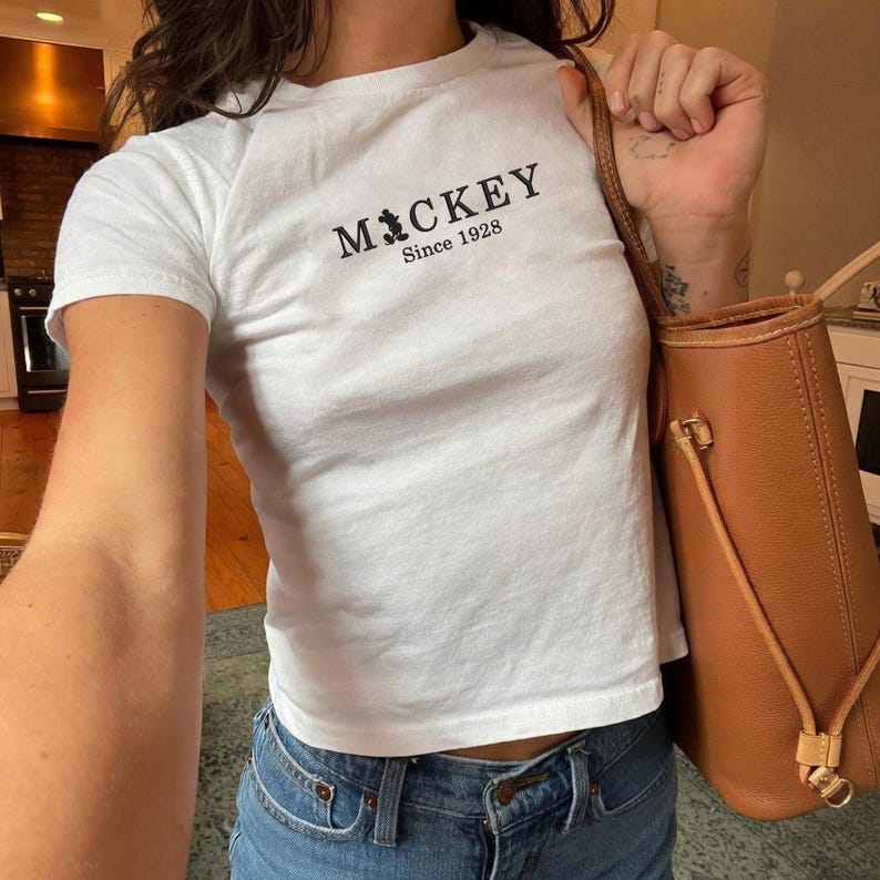May include: A white short-sleeved t-shirt featuring the black text MICKEY with a small silhouette of Mickey Mouse replacing the letter I, and the text Since 1928 underneath. The shirt is paired with blue denim jeans and a tan leather tote bag.