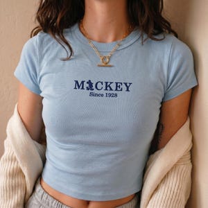 May include: A light blue short-sleeved t-shirt featuring dark blue embroidered text that reads Mickey Since 1928, with the letter i replaced by a silhouette of Mickey Mouse. The shirt has a crew neckline and is worn with a gold-toned toggle necklace.