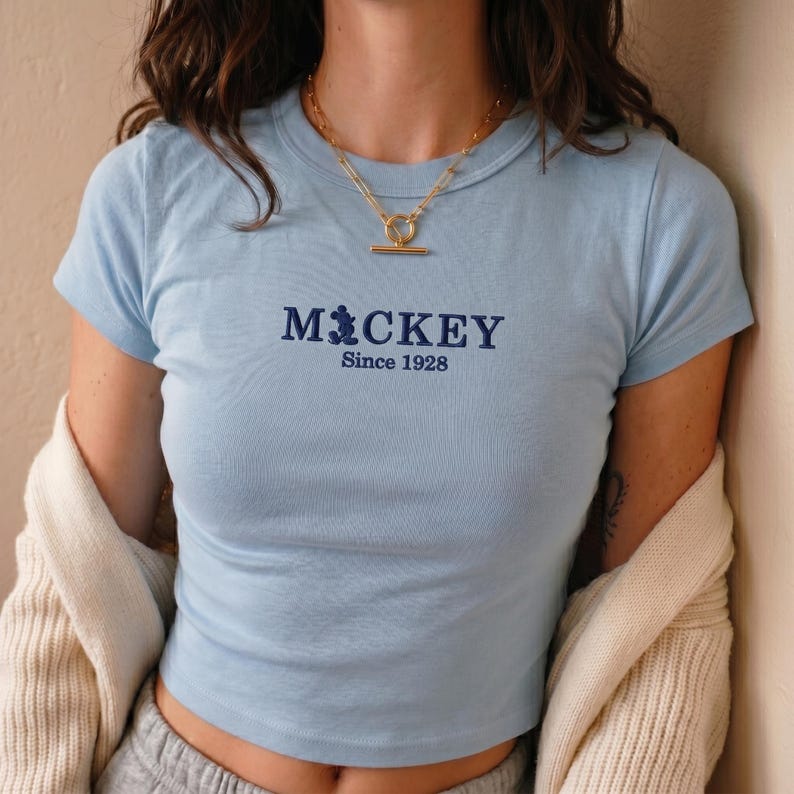 May include: A light blue short-sleeved t-shirt featuring dark blue embroidered text that reads Mickey Since 1928, with the letter i replaced by a silhouette of Mickey Mouse. The shirt has a crew neckline and is worn with a gold-toned toggle necklace.