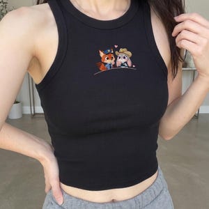 May include: Black ribbed tank top featuring an embroidered design of a fox and a bunny, each wearing a hat. The tank top has a round neckline and a fitted silhouette. The design includes a small pink heart above the characters.