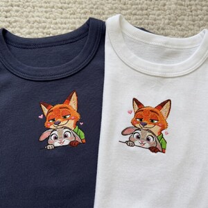 May include: Two short-sleeved t-shirts, one navy blue and one white. Each shirt features an embroidered design of a fox and a rabbit, with pink heart accents. The fox is orange and the rabbit is gray.