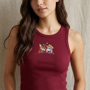 May include: Burgundy ribbed tank top with an embroidered design featuring two cartoon characters. One is a fox wearing a police uniform, and the other is a bunny wearing a cowboy hat. The tank top has a scoop neck and a fitted silhouette.
