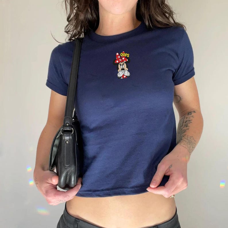 Op de afbeelding: Een marineblauw T-shirt met korte mouwen en een kleine geborduurde afbeelding van Minnie Mouse met een rood-witte stippenstrik, die haar mond met haar handen bedekt, vergezeld van de gele tekst OOPS boven haar hoofd.