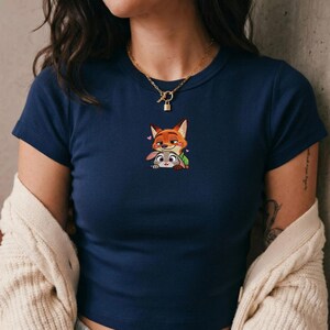 May include: Navy blue short-sleeve t-shirt featuring an embroidered image of Nick Wilde and Judy Hopps from Zootopia. The design includes a fox and a bunny with hearts. The shirt has a round neckline.
