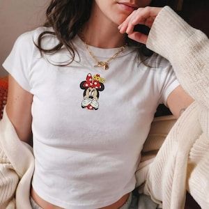Op de afbeelding: Een wit T-shirt met korte mouwen voorzien van een kleine afbeelding van Minnie Mouse die haar mond met haar handen bedekt, een rood-witte stippenstrik draagt, met de gele tekst OOPS boven haar hoofd en een gouden ketting.