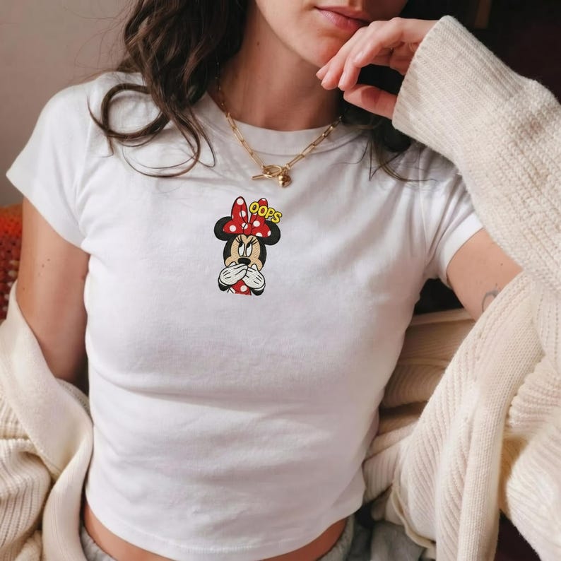 Op de afbeelding: Een wit T-shirt met korte mouwen voorzien van een kleine afbeelding van Minnie Mouse die haar mond met haar handen bedekt, een rood-witte stippenstrik draagt, met de gele tekst OOPS boven haar hoofd en een gouden ketting.