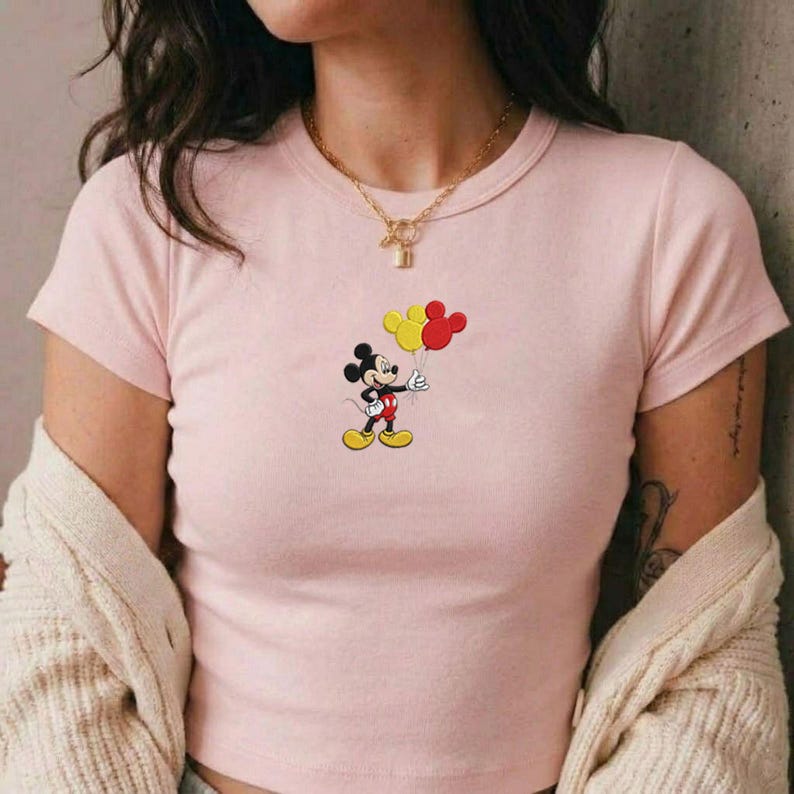 Op de afbeelding: Een lichtroze T-shirt met korte mouwen en een afbeelding van Mickey Mouse die twee ballonnen vasthoudt, &eacute;&eacute;n gele en &eacute;&eacute;n rode, in de vorm van Mickey-hoofden. Het shirt heeft een ronde hals en wordt gedragen met een cr&egrave;me vest over de schouders.