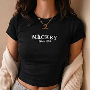 May include: A black short-sleeved t-shirt featuring white embroidered text that reads MICKEY with a small silhouette of the character replacing the letter I, followed by the text Since 1928. A gold-toned chain necklace with a toggle clasp.
