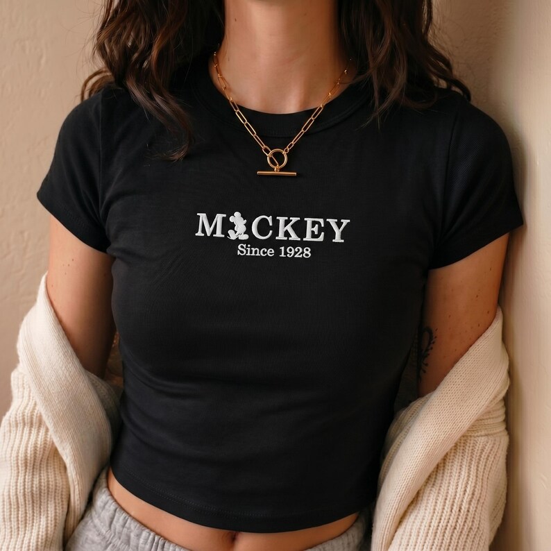 May include: A black short-sleeved t-shirt featuring white embroidered text that reads MICKEY with a small silhouette of the character replacing the letter I, followed by the text Since 1928. A gold-toned chain necklace with a toggle clasp.