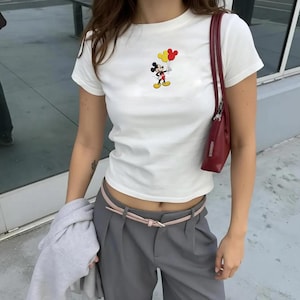 Op de afbeelding: Een wit T-shirt met korte mouwen en een kleine afbeelding van Mickey Mouse die drie gele en rode ballonnen vasthoudt. Het shirt wordt gecombineerd met een grijze broek met een dunne roze riem en een donkerrode schoudertas met smalle band.